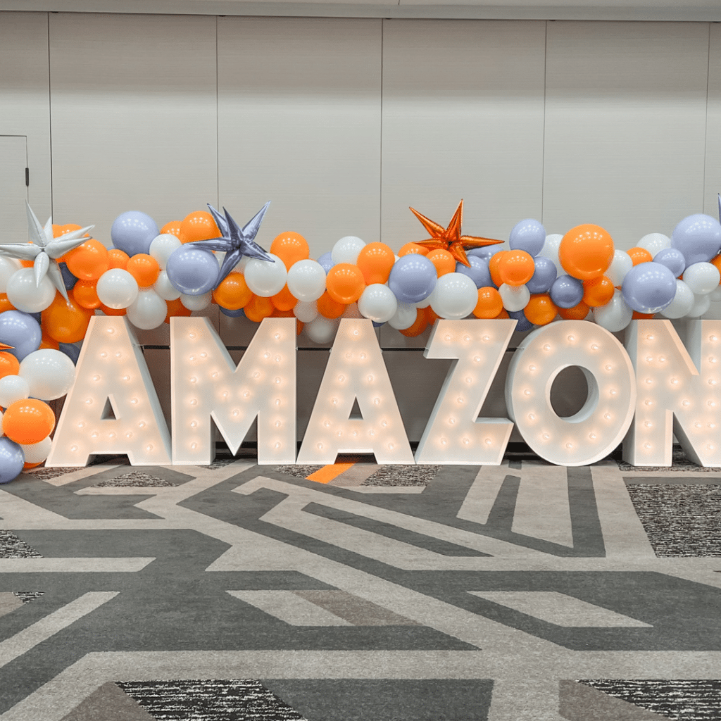 Marquee Letters spelling out AMAZON beneath orange, white, and gray balloons