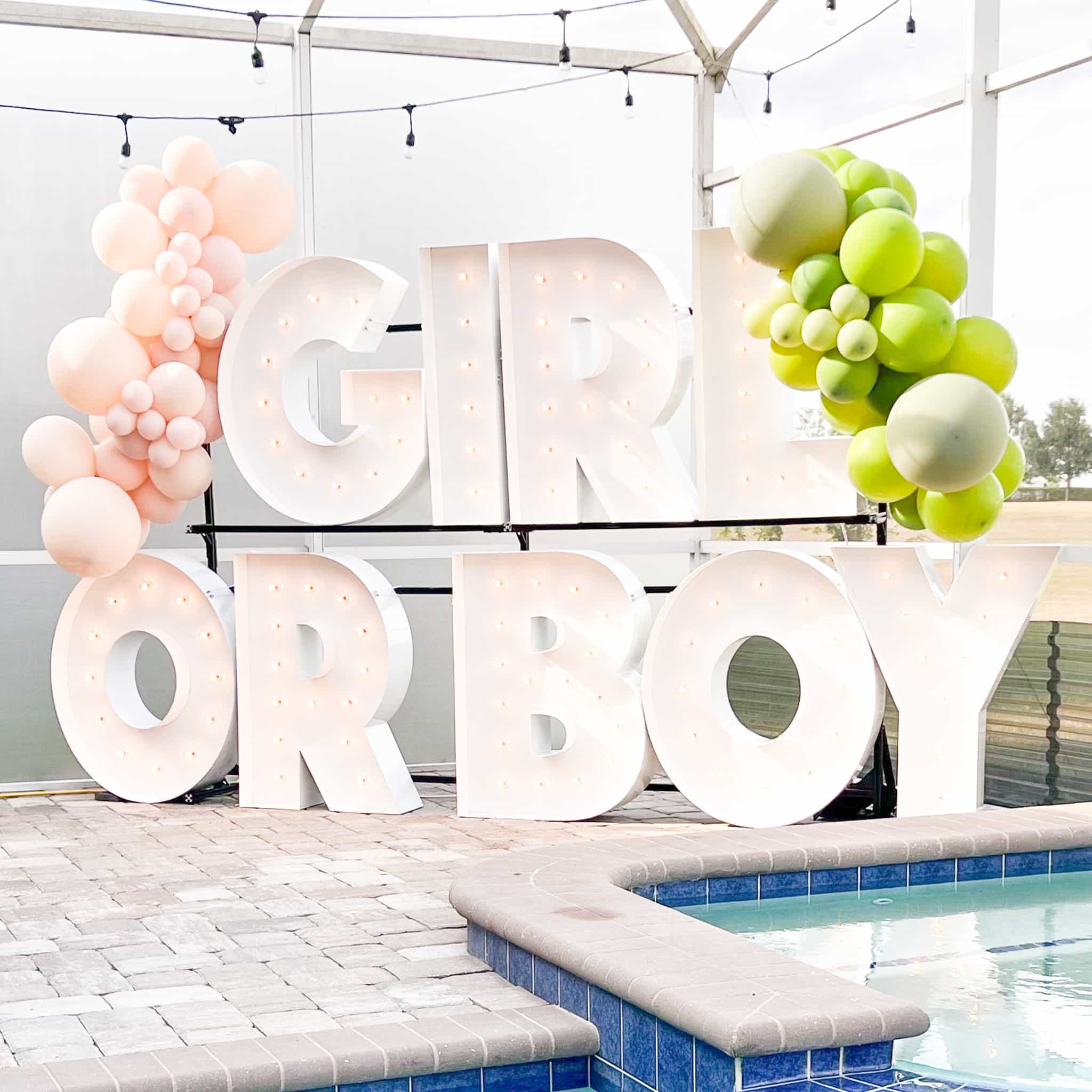 Marquee letter display in a backyard that reads "GIRL OR BOY" with orange and green balloons
