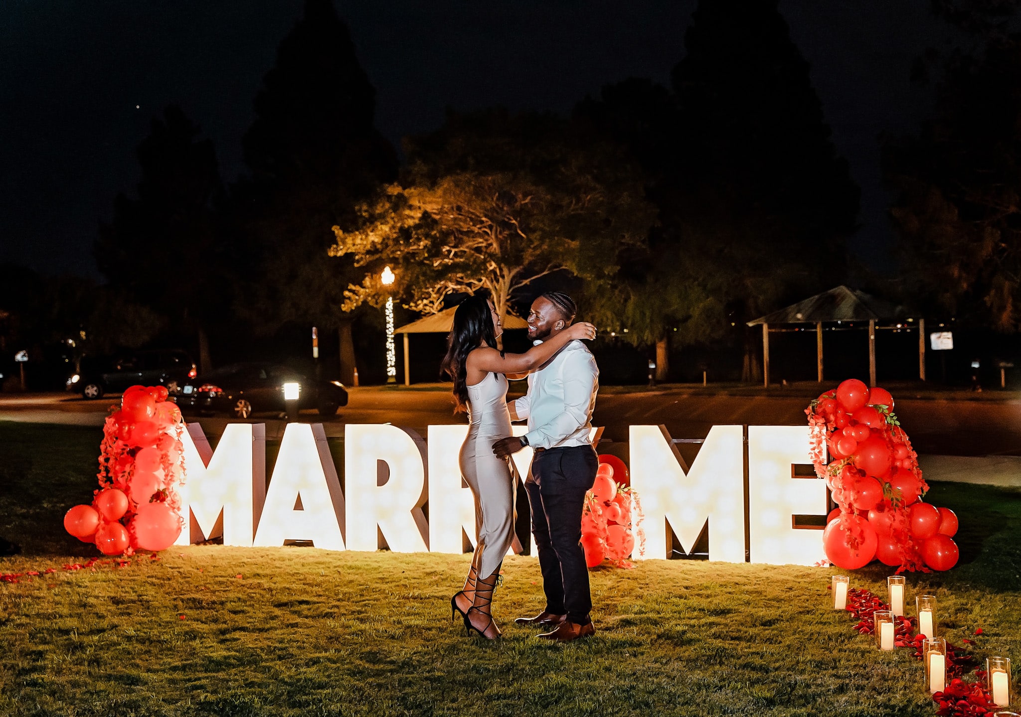 A man and a woman embracing in from of a light up letter display that reads "MARRY ME"