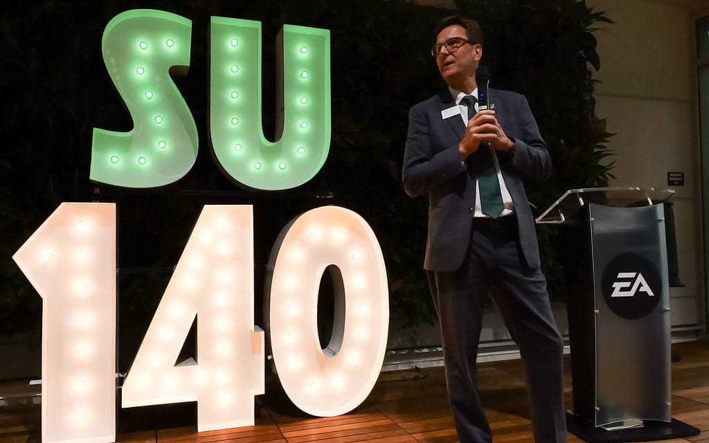 Speaker at a podium for EA Sports in front of marquee light up letters that read SU 140
