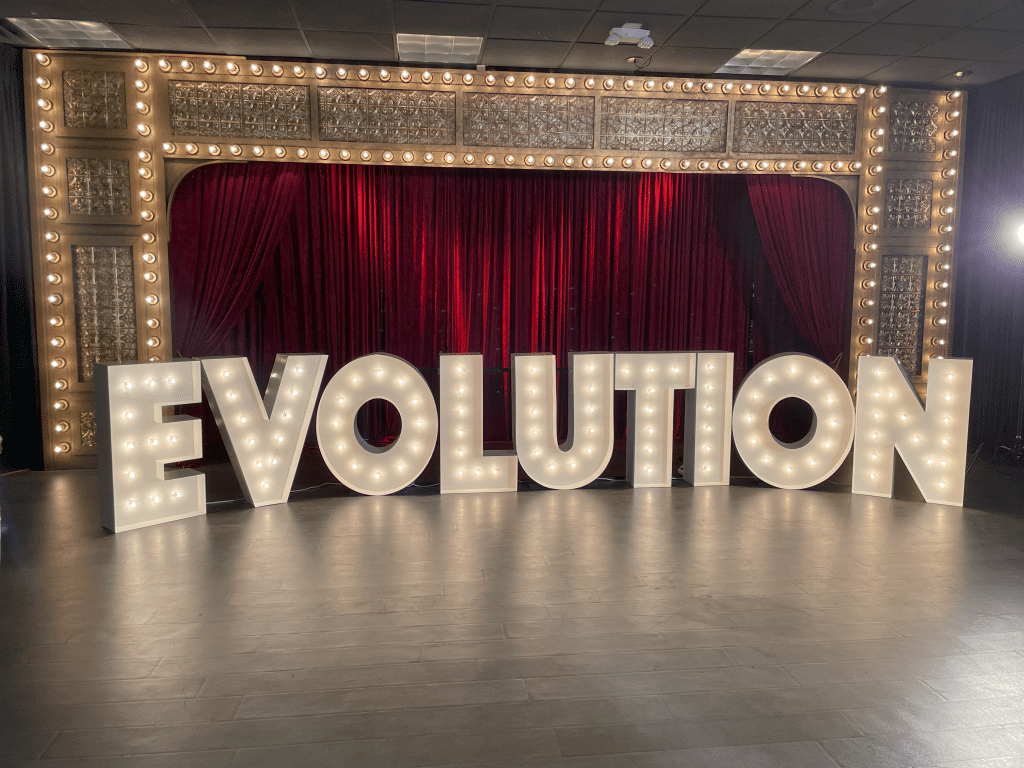 Light up marquee letters spelling out EVOLUTION in front of a theater curtain