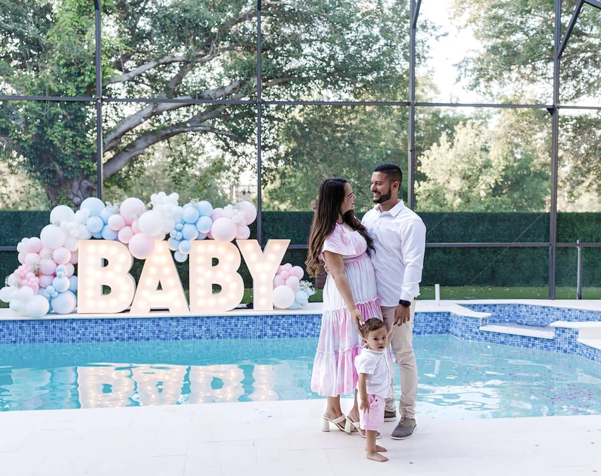 The word baby lit up in marquee letters near a pool. The word is surrounded by pink and blue balloons while the family stands in the front.