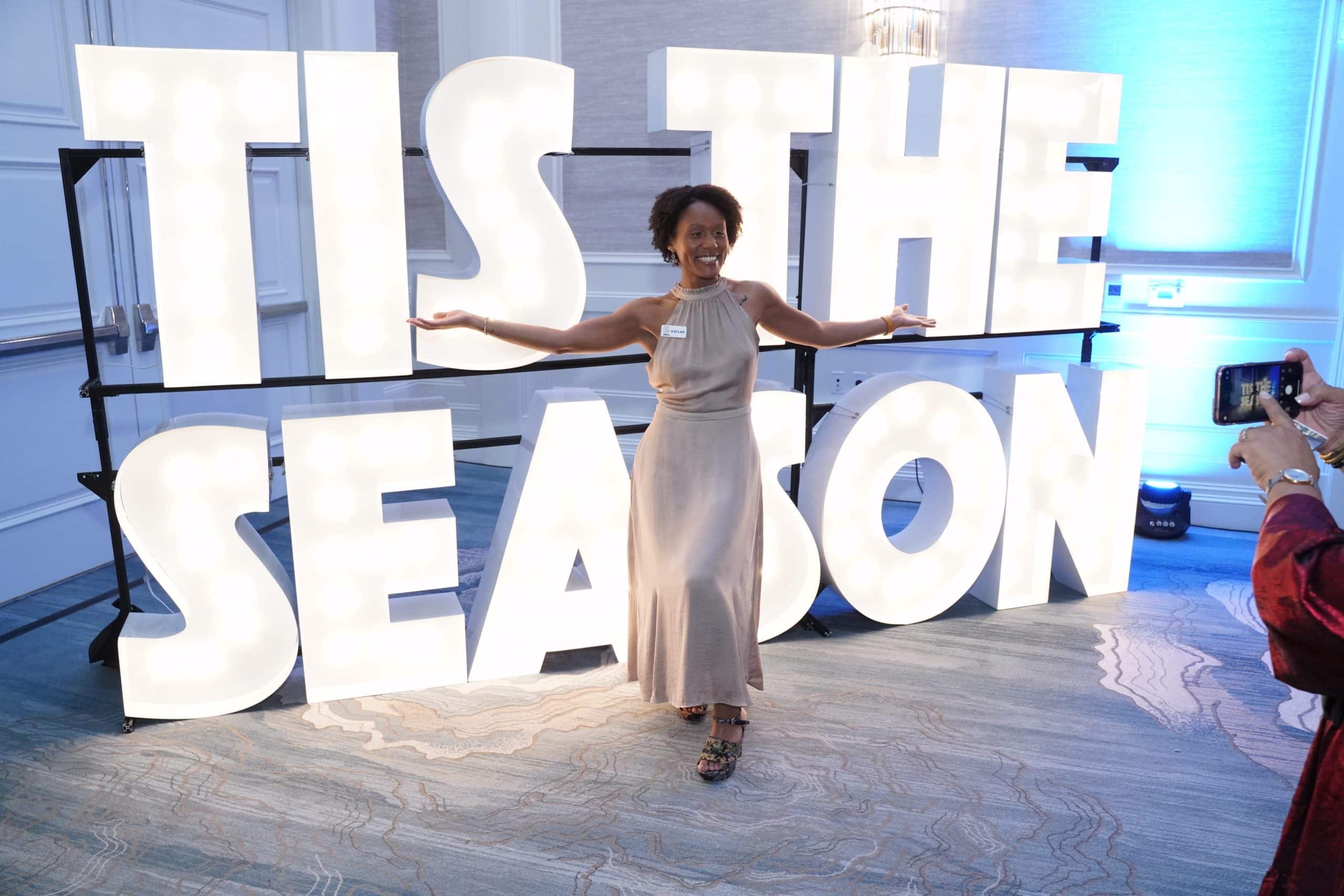 Event-Lit business owner in front of a two-tier marquee letter display that reads "TIS THE SEASON"