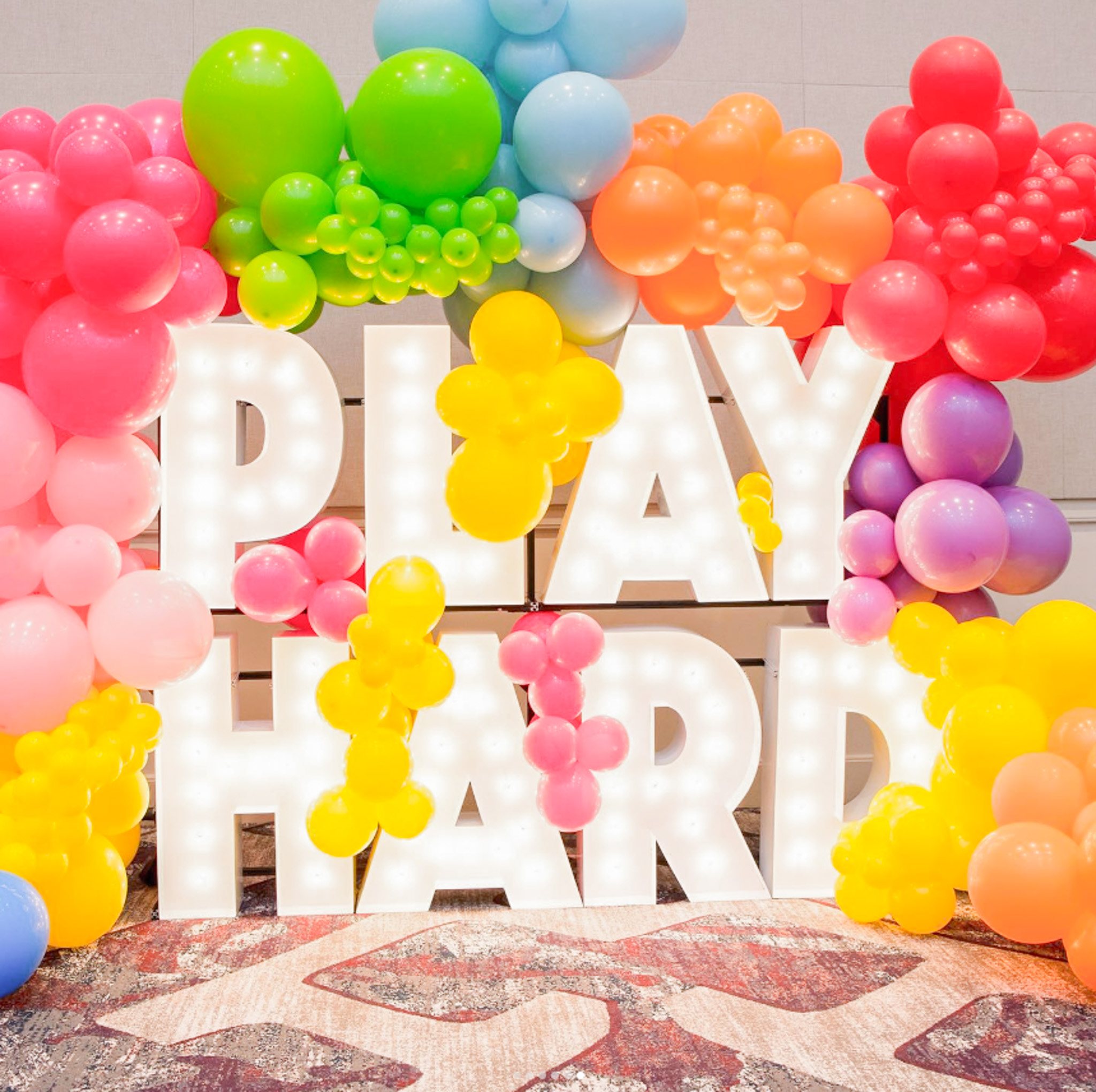 Play Hard spelled out in marquee letters. There are blue, yellow, pink, green, orange, and red balloons surrounding it.